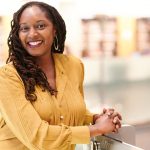 A woman with dreadlocks and a bright smile, wearing a mustard yellow blouse, standing in a modern library with bookshelves in the background.